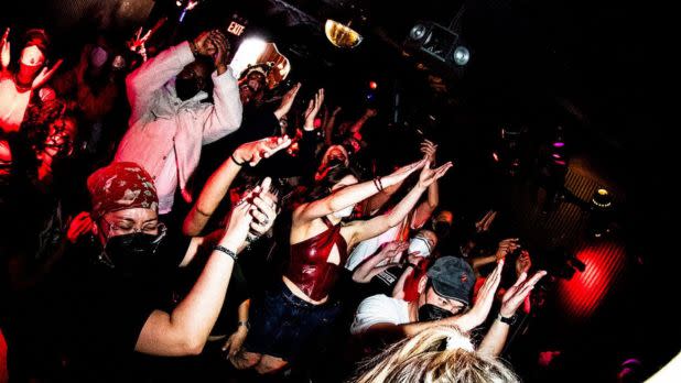 A crowd of people dances with their hands in the air at a packed nightclub or concert venue, illuminated by red and purple stage lighting, creating a high-energy, celebratory atmosphere.