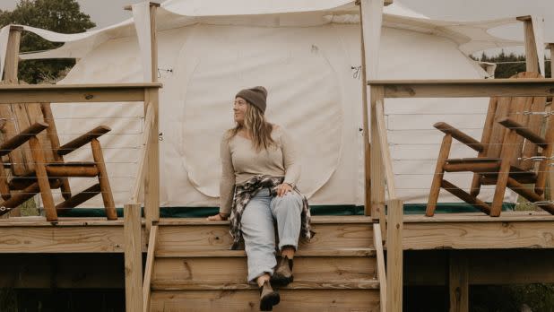 Woman in a beanie sits on wooden steps outside a canvas tent, wearing casual layers; rustic glamping setting evokes a calm, cozy mood.
