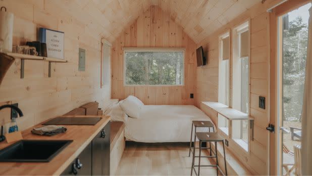 Cozy wood-paneled tiny home interior with a bed, compact kitchen, stools, and sunlit windows overlooking trees; a warm, peaceful retreat.