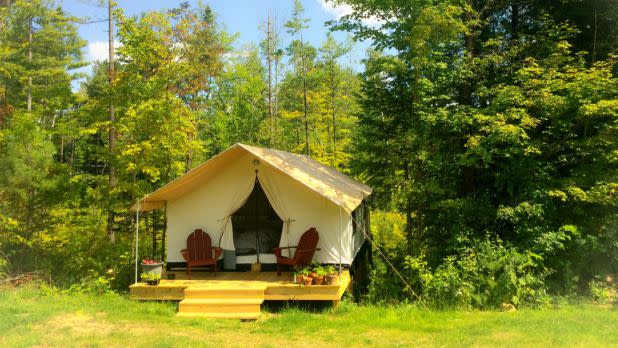 A cozy canvas tent cabin with open flaps, two red Adirondack chairs, and flower boxes sits on a sunny porch in a lush green forest.