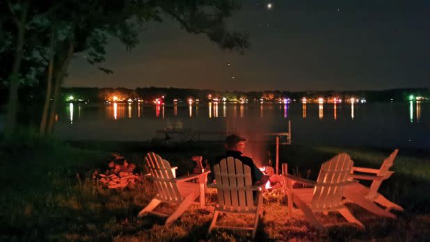 Three Adirondack chairs face a glowing lakeside fire at night, with colorful town lights reflecting on the water beneath a starry sky.