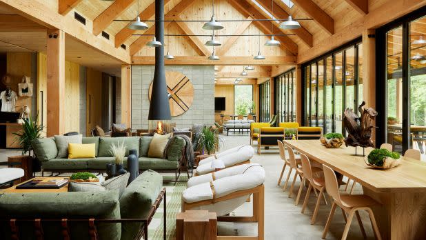 Open-plan timber lodge interior with exposed beams, cozy seating, dining table, plants, and expansive windows creating a warm, inviting atmosphere.