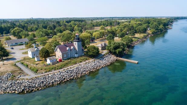 Aerial view of a historic lakeside lighthouse, homes, rocky shoreline, and dock beside calm, clear blue-green water on a sunny day.