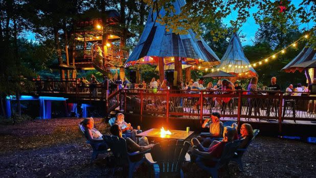 Nighttime campsite with guests gathered around a glowing fire table, colorful tents and string lights on a wooden deck, creating a lively, cozy mood.