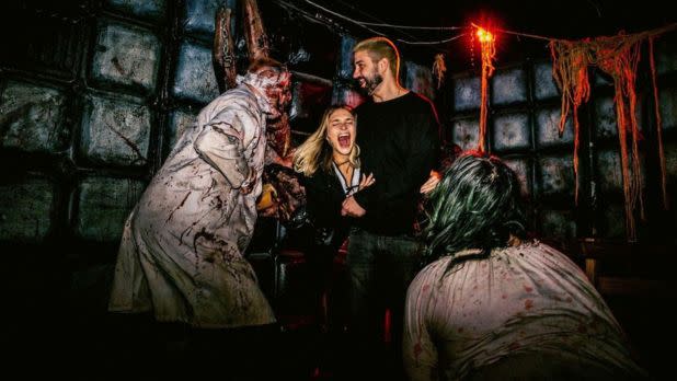 In a dim, blood-streaked room, a woman laughs beside a man as grimy, zombie-like figures loom nearby, creating a playful horror scene.
