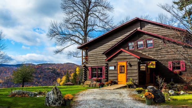 A rustic timber lodge with red trim and a bright yellow door stands beside a gravel path, framed by autumn hills, green lawn, and cloudy blue skies.