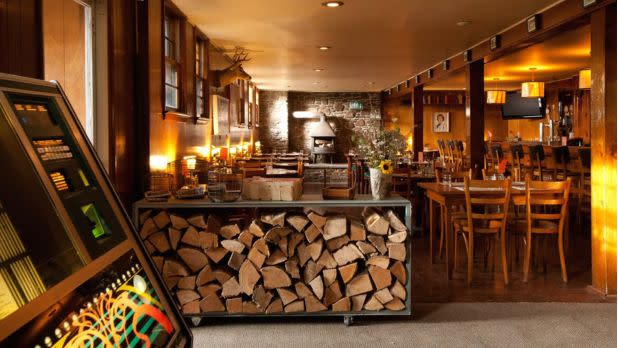 A cozy rustic restaurant interior glows with amber lighting, featuring a wood-fired hearth, stacked logs, wooden tables, and a menu kiosk.