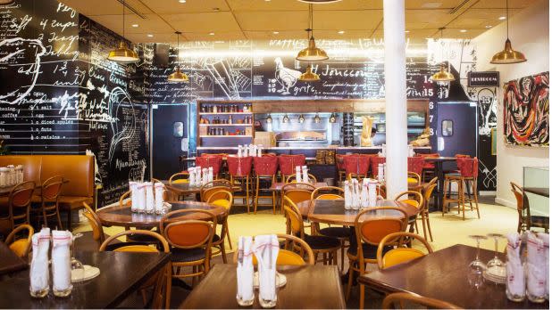 A warmly lit restaurant interior features wooden tables, orange chairs, red stools, and chalkboard walls covered in handwritten notes, creating a lively, eclectic atmosphere.