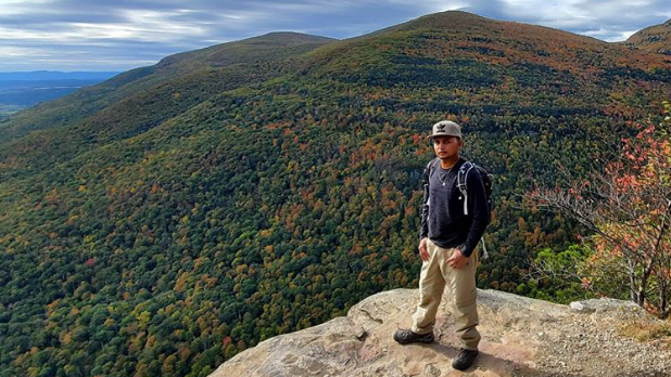 Official I LOVE NY Leaf Peeper Feroz standing in front of a mountain with fall foliage