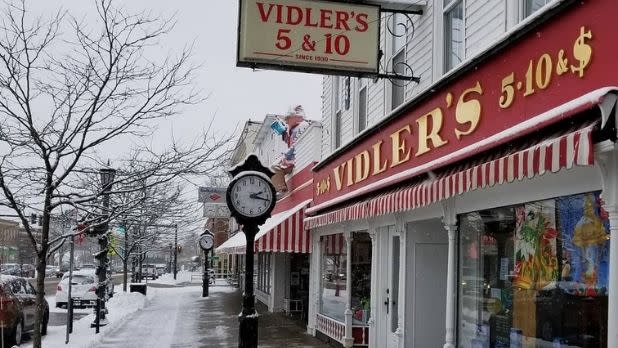 A snow-covered street view of Vidler's red and white colored 5 & 10 storefront with a red and white pinstriped awning and a large clock post standing in front of it
