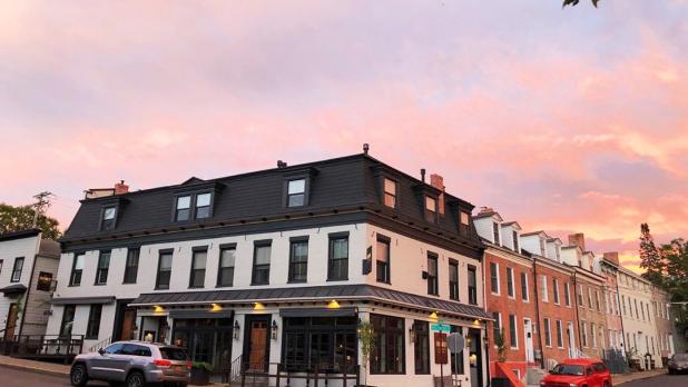 The white exterior of WM Farmers and Sons with a grey car parked on the left of the corner and a red car on the right of the corner with the sun setting outside the frame creating a cotton candy sky