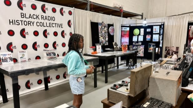 A young girl observes vintage radio equipment at the WUFO Black Radio Historic Collective exhibit. Display tables and informational boards surround her.