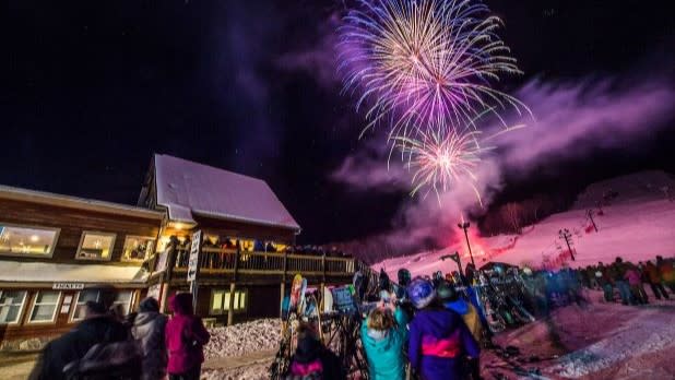 People in winter clothing watch as a vibrant fireworks display illuminates the night sky over West Mountain during the ski resort's annual Fire on the Mountain event.