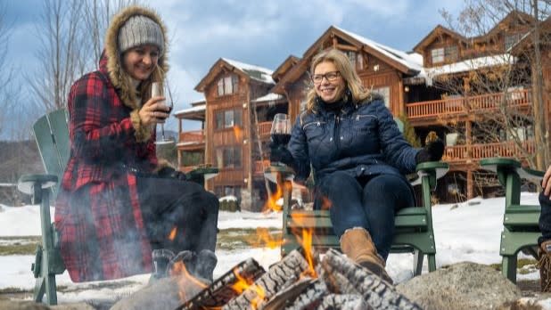 Two women sit by a warm fire, holding wine glasses, in green chairs at Whiteface Lodge. They are dressed in winter clothing, with snowy wooden lodges in the background.