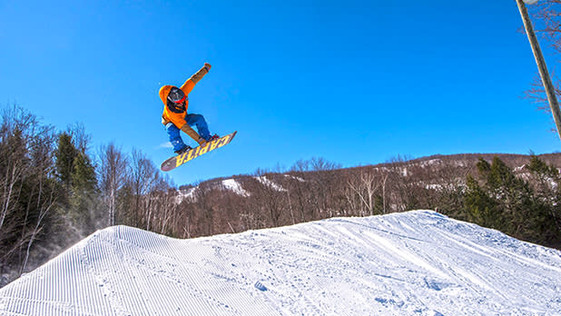 A snowboarder in a bright orange jacket and blue pants is flying through the air on a snowboard along a snowy slope at Windham Mountain in the catskills