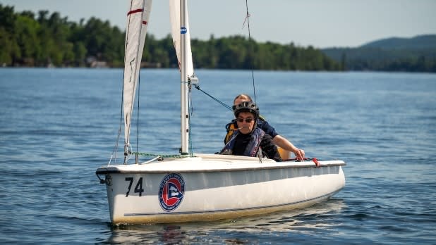 A person uses a sip 'n' puff system to steer a small sailboat on Lake George with Y-Knot Sailing. The sun is bright, and pine tree-covered hills are visible on the horizon.