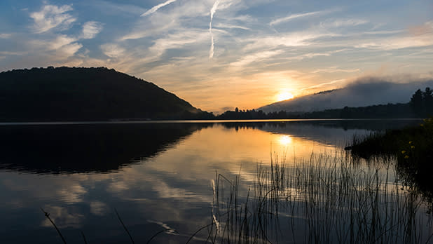 The sun rises behind a hill overlooking a lake at the Red House Area of Allegany State Park