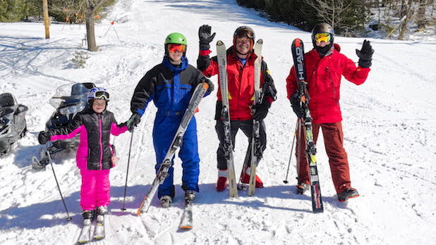 Four people in ski suits pose for a photo at the bottom of a slope at Holiday Mountain