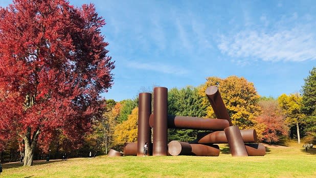 A metal sculpture comprised of oversized cylindrical rods stands on the grassy field of Storm King Art Center with trees bursting in fall colors in the background
