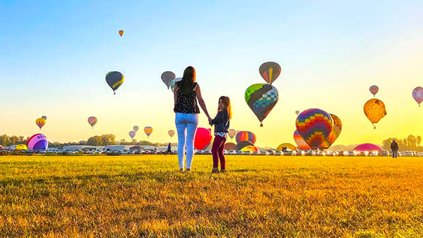 A woman and girl standing in a yellow grassy field  watching a group of hot air balloons landing as the sun sets