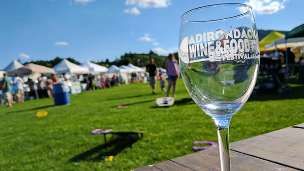 A wine glass stands on a wooden table with a green lawn and tents in the background at the Adirondack Wine & Food Festival