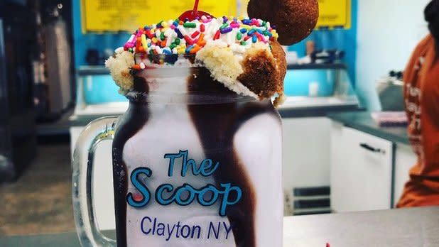 A milkshake with hot fudge, whipped cream, sprinkles, and a cherry in a mason jar that says "The Scoop, Clayton NY"