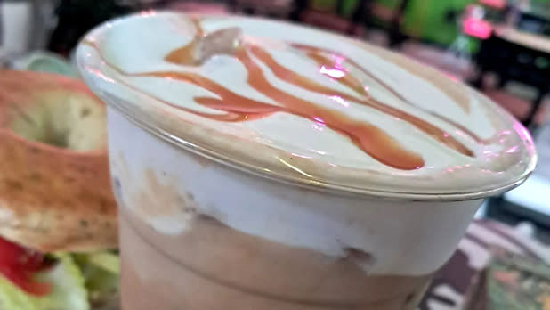 A plastic cup filled with iced coffee topped with cream and caramel drizzle, next to a bagel sandwich.