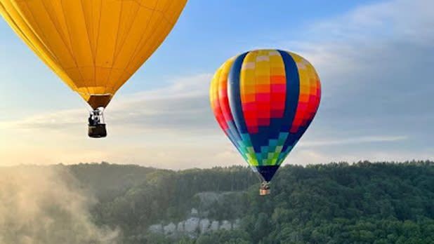 Two hot air balloons soar through the blue skies over the green forests at Letchworth