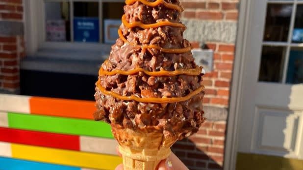 Close-up of a hand holding a waffle cone with chocolate-dipped rocky road ice cream, drizzled with caramel. Background shows colorful bench and brick wall.