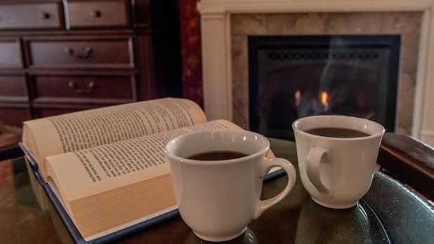Two steaming cups of tea sit on a table next to an open book in front of a fire place