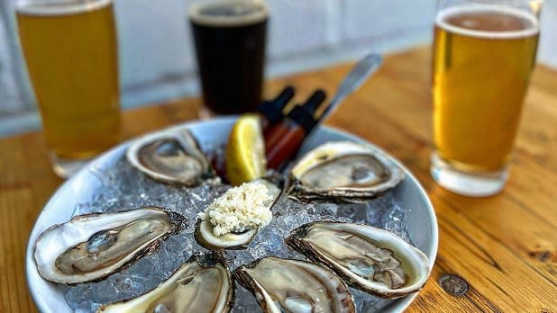 A bowl of fresh oysters on ice sits on a table with three glasses of beer