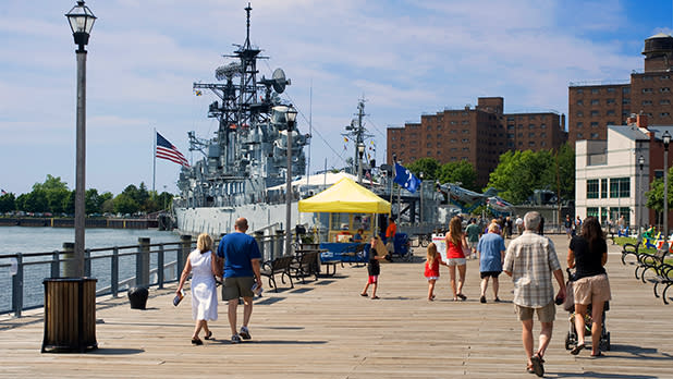 People walking along the boardwalk in Canalside Buffalo in summer with a large ship docked to the marina