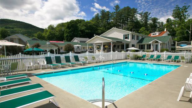 Sunny outdoor scene with a clear blue pool, surrounded by green lounge chairs and a white fence. A white house and lush trees in the background. Relaxing ambiance.