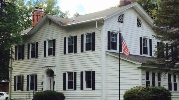 White colonial-style house with black shutters, a red brick chimney, and an American flag. The setting is calm with trees and bushes nearby.