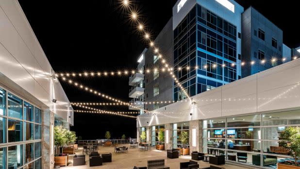Modern outdoor courtyard at night with string lights, surrounded by glass buildings. Furnished with black chairs and tables, creating a cozy, urban vibe.