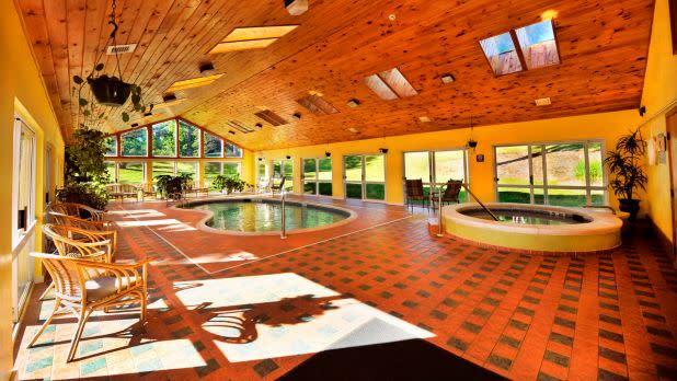 Sunny indoor pool area with a wooden ceiling and yellow walls. Large windows let in sunlight, illuminating the pool, hot tub, and wicker chairs. Cozy and inviting atmosphere.