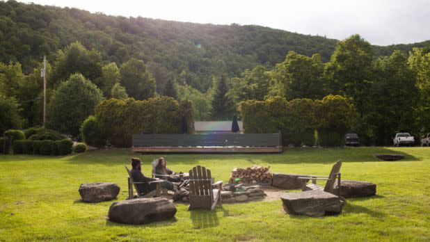 Two people relax in Adirondack chairs around a fire pit, surrounded by lush greenery and hills. The scene is peaceful and inviting.
