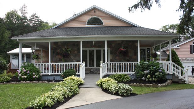 A charming single-story house with a stone facade and a wide, white-railed porch. Lush greenery and blooming flowers line the path, creating a welcoming feel.