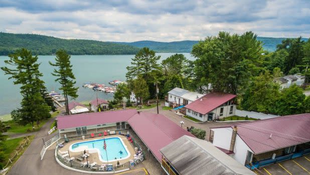 Aerial view of a lakeside resort with a red-roofed building surrounding a pool. The lake, boats, and lush trees create a serene, inviting atmosphere.