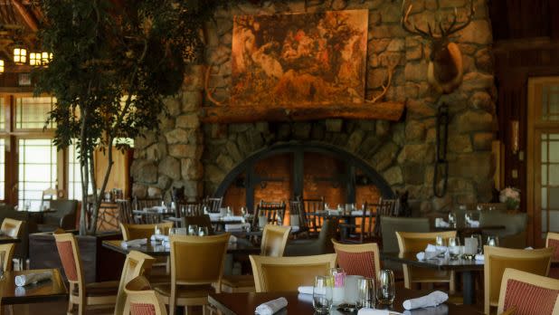 Cozy restaurant interior with stone fireplace, animal mount on the wall, and tables set for dining. Warm and rustic atmosphere with natural light.