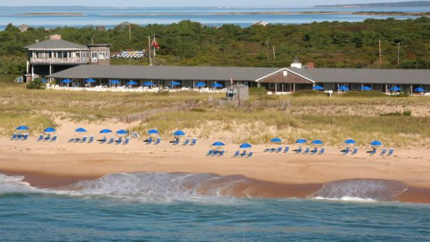 A serene beach scene with rows of blue umbrellas and lounge chairs on golden sand. A long beachside building is surrounded by lush greenery and distant waters.