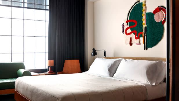Modern hotel room with a large bed featuring white linens, abstract wall art, and a cozy green sofa. Large window with black curtains adds natural light.