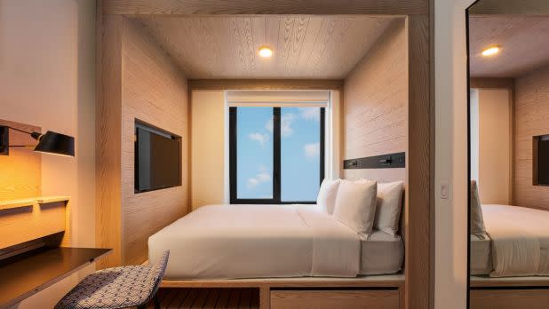 A cozy, modern hotel room features a neatly made bed beside a large window showcasing a blue sky with clouds. Warm wood tones, a small desk, and a sleek TV create a serene, minimalist vibe.
