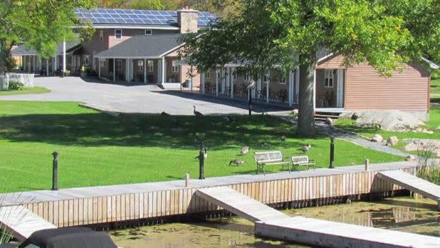 A tranquil lakeside scene with a dock in the foreground, lush green grass, and geese. In the background, a house with solar panels is partially shaded by trees.