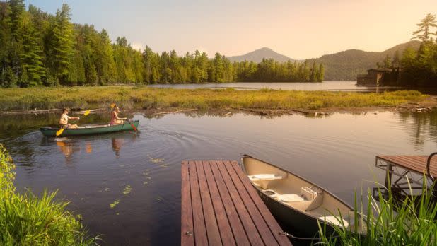 Two people paddle a canoe on a serene lake, surrounded by lush trees and mountains at sunset. A wooden dock and an empty canoe await in the foreground.