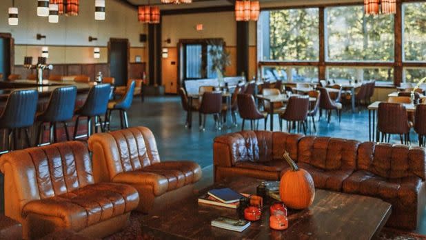 Cozy lounge with brown leather sofas and a wooden table featuring a pumpkin and books. Warm lighting and large windows offer a welcoming autumn vibe.