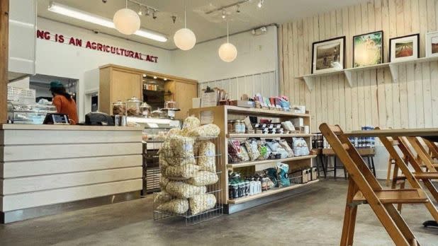 Cozy café interior with wooden shelves displaying snacks, a counter with a staff member, pendant lights, and framed art. Warm, inviting atmosphere.
