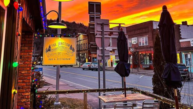 A vibrant sunset casts an orange glow over a street and The Junction Bar & Restaurant sign. Tables with closed umbrellas and colorful lights create a cozy atmosphere.