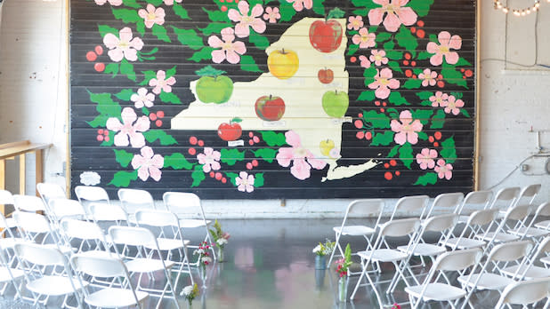 View from the aisle of a wedding ceremony setup at Nine Pin Ciderworks with mural of New York State with pink flowers and apples