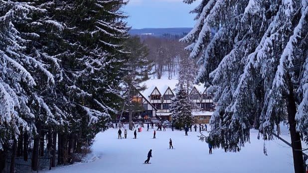 Snowy winter scene with people skiing near an alpine lodge at Peek'n Peak Resort. Tall snow-laden trees frame the view, creating a serene and festive atmosphere.